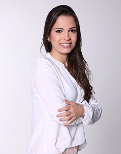 foto profissional da psicóloga milene de camisa de manga comprida, branca, de braços cruzados, e calça na cor salmão